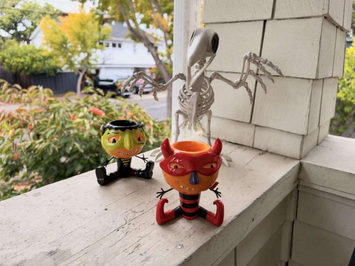 Two ceramic Halloween candle holders, one a Frankenstein-style figure and one a red devil, sit on a porch railing beside a skeleton bird decoration, with a leafy garden and street behind them.