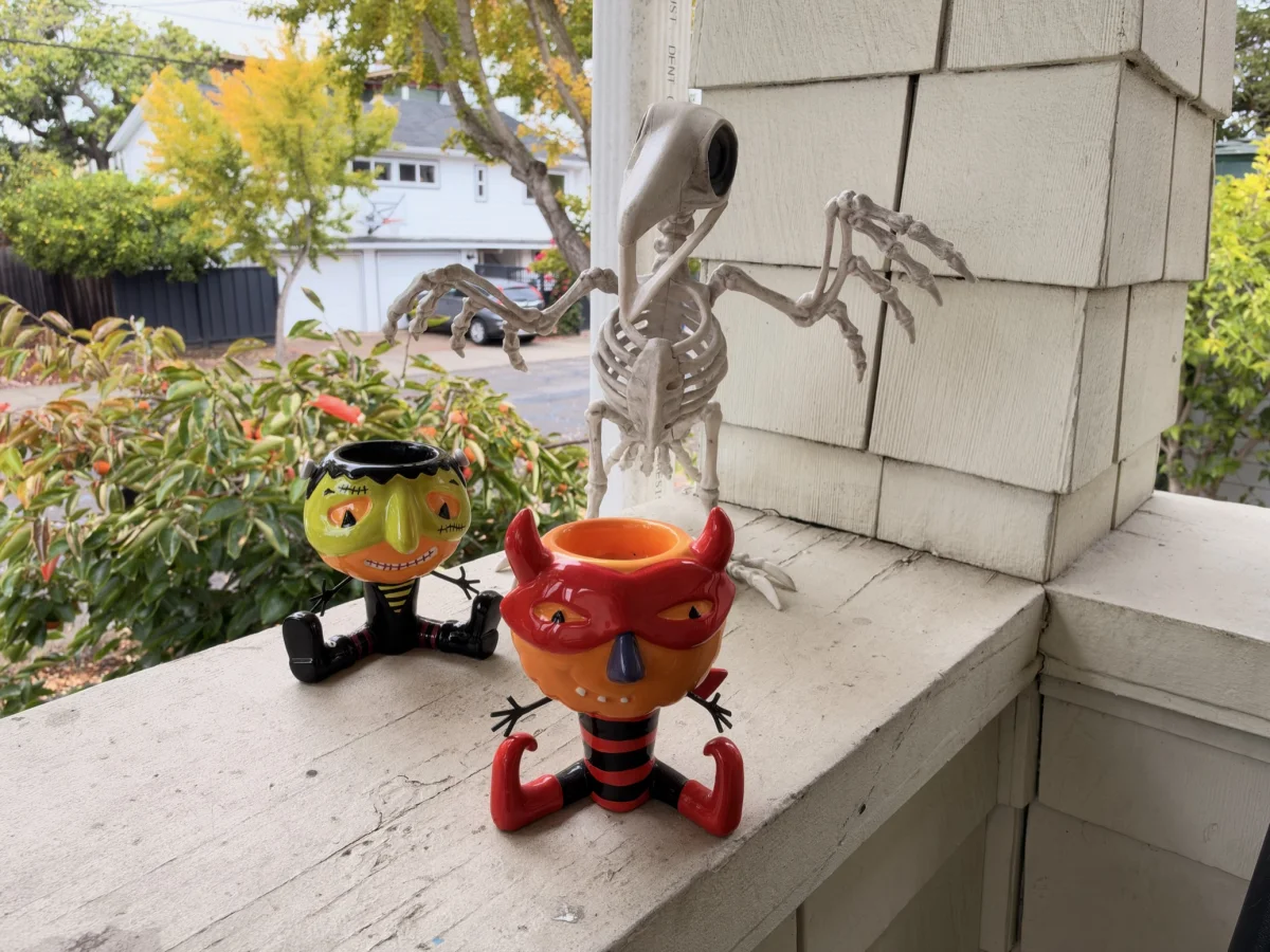 Two ceramic Halloween pumpkin-head candy bowls, one dressed as Frankenstein and one as a red devil, sit on a porch railing beside a plastic skeleton bird. Autumn trees and a neighbor's house are blurred behind them.