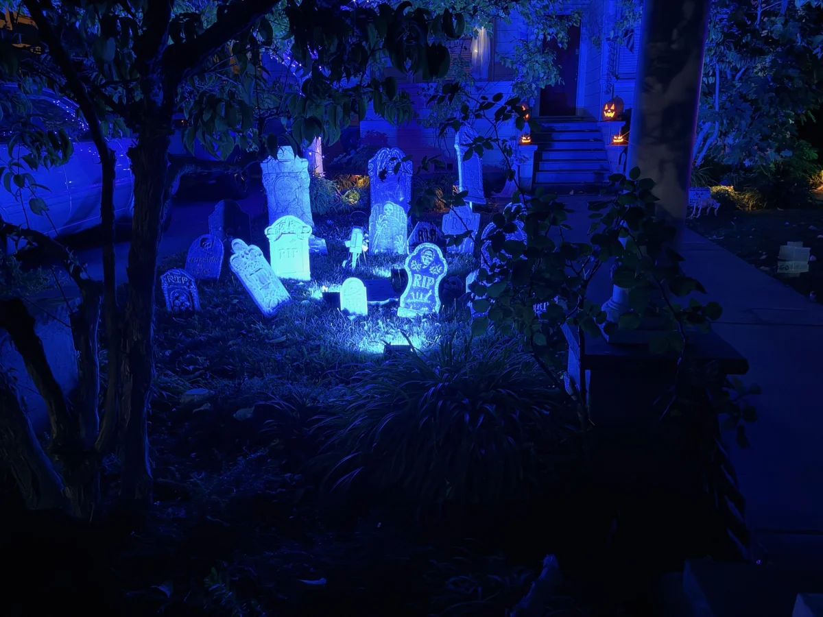 A front yard at night lit in eerie blue, filled with foam Halloween tombstones marked RIP and skulls. In the background, glowing jack-o'-lanterns sit on the steps of a house, framed by dark tree branches and foliage.