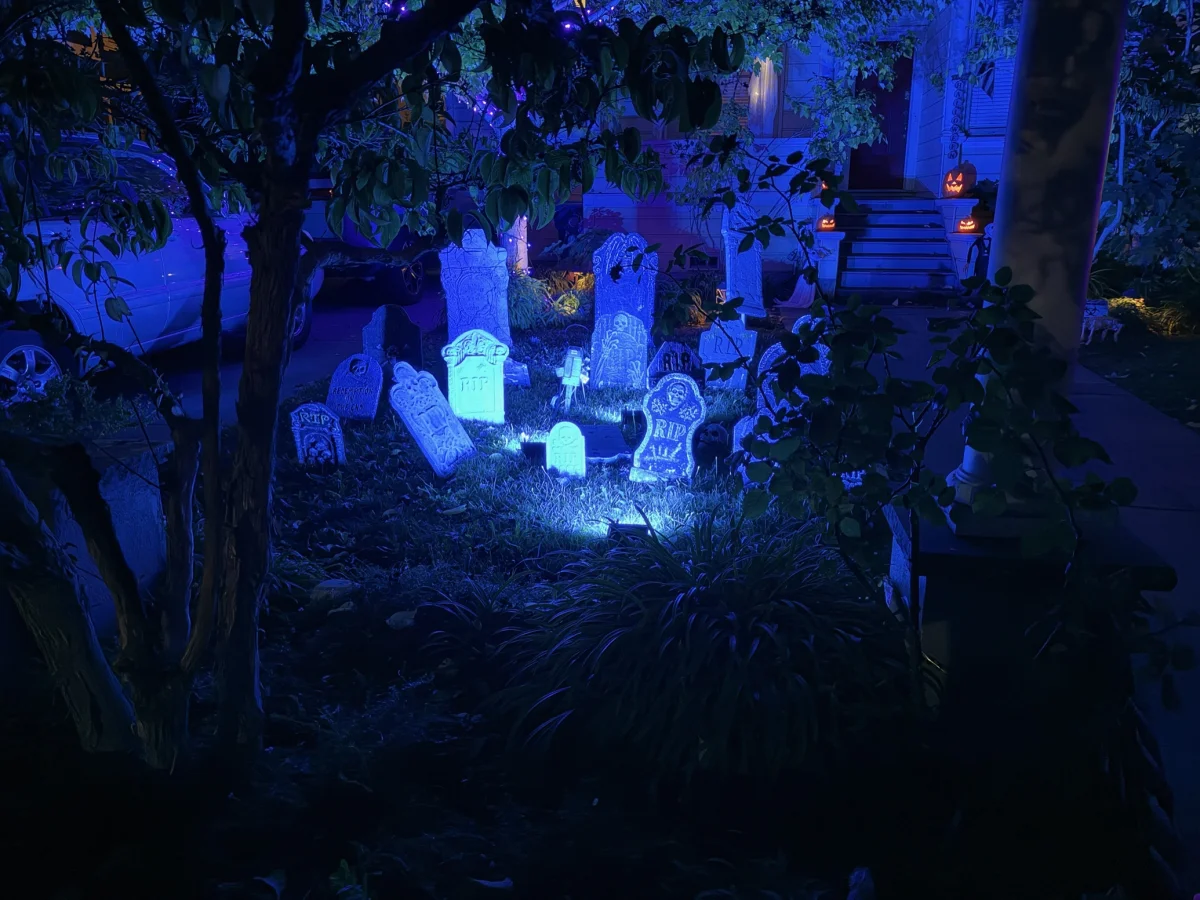 A front yard decorated for Halloween at night, with foam tombstones lit by blue light. Glowing jack-o'-lanterns sit on the porch steps of a house in the background, and a parked car is visible at left.