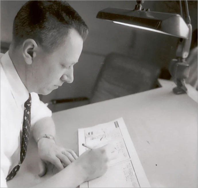 A black-and-white photo of Charles Schulz sitting at a drafting table, drawing Peanuts with a pencil under a desk lamp.