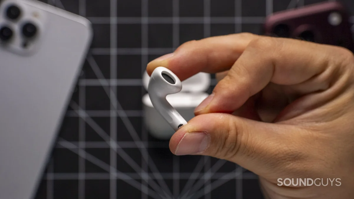Close-up of a white Apple AirPod held beside its charging case and two smartphones