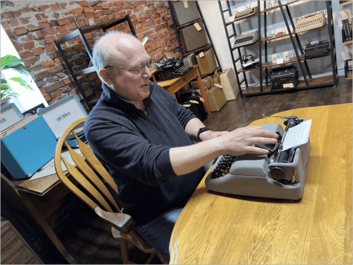Glenn sitting at a wooden table in a typewriter store, typing on a gray manual typewriter with shelves of vintage typewriters in the background.
