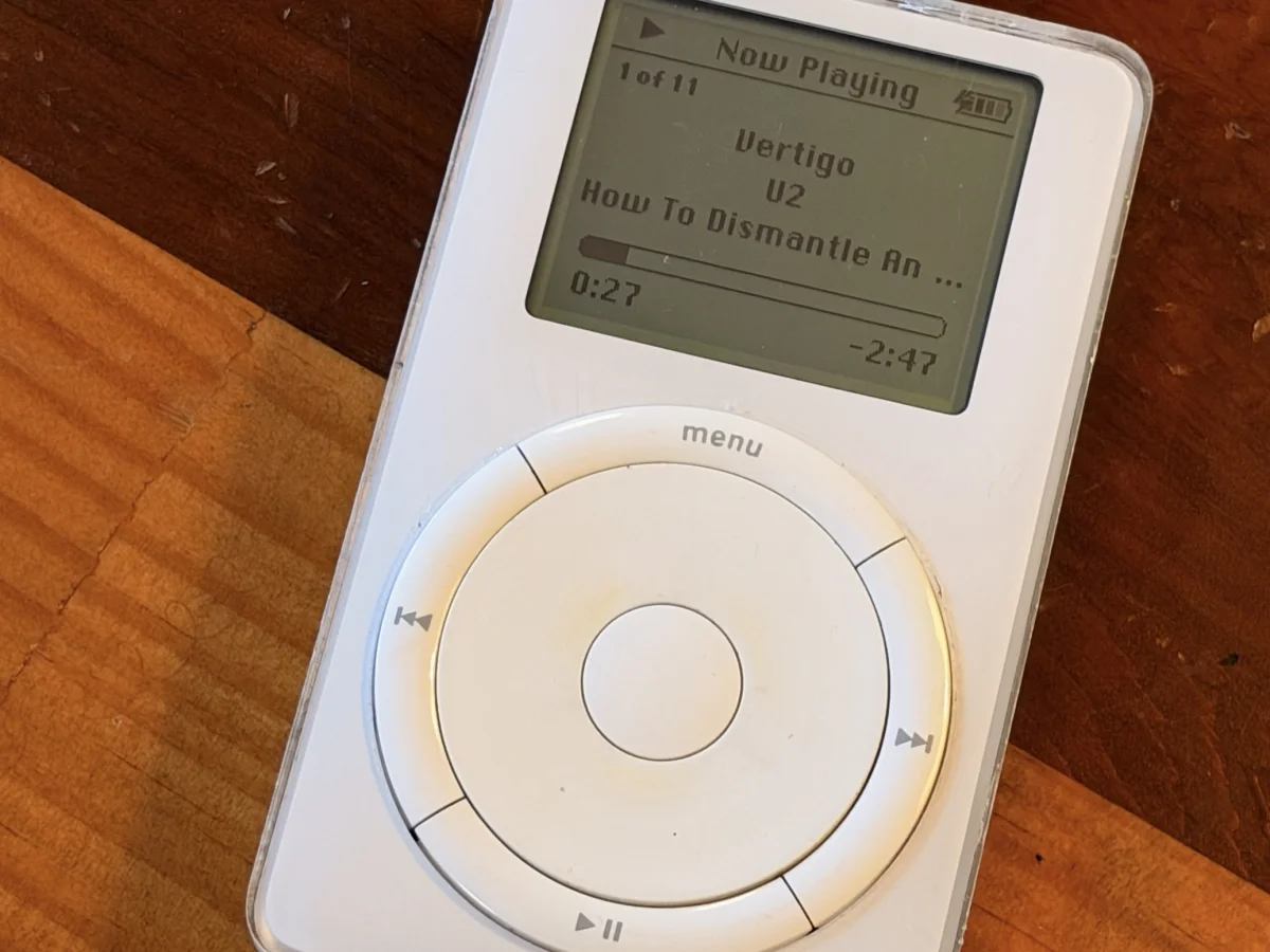 A first-generation iPod with a scroll wheel sits on a wooden table. Its screen shows Now Playing: "Vertigo" by U2 from How To Dismantle An Atomic Bomb, 0:27 elapsed with 2:47 remaining.