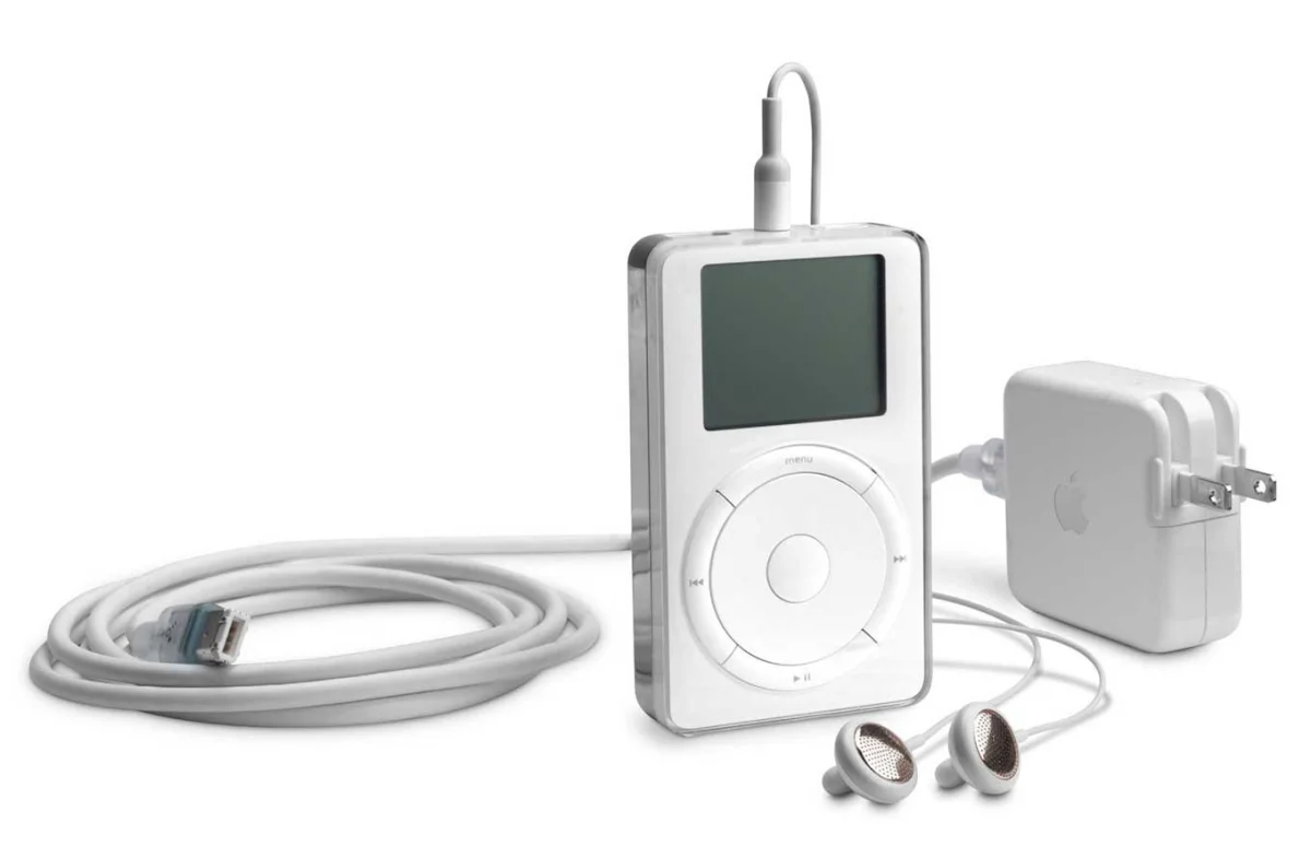 The original white Apple iPod with its monochrome screen and scroll wheel, shown with a coiled FireWire cable, white earbuds, a headphone plug at the top, and a white Apple power adapter with folding prongs.