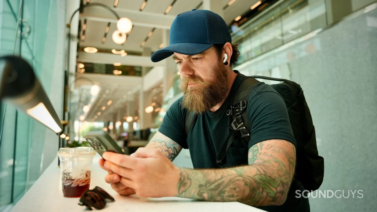 A person wearing AirPods Pro 3 earbuds while using a smartphone with a backpack and iced coffee nearby.