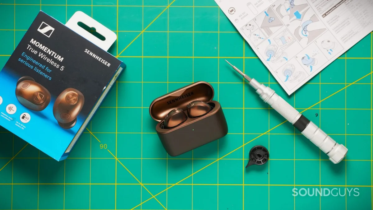 The bronze Sennheiser Momentum True Wireless 5 earbuds sit in their open charging case beside the retail box, screwdriver, and instructions.